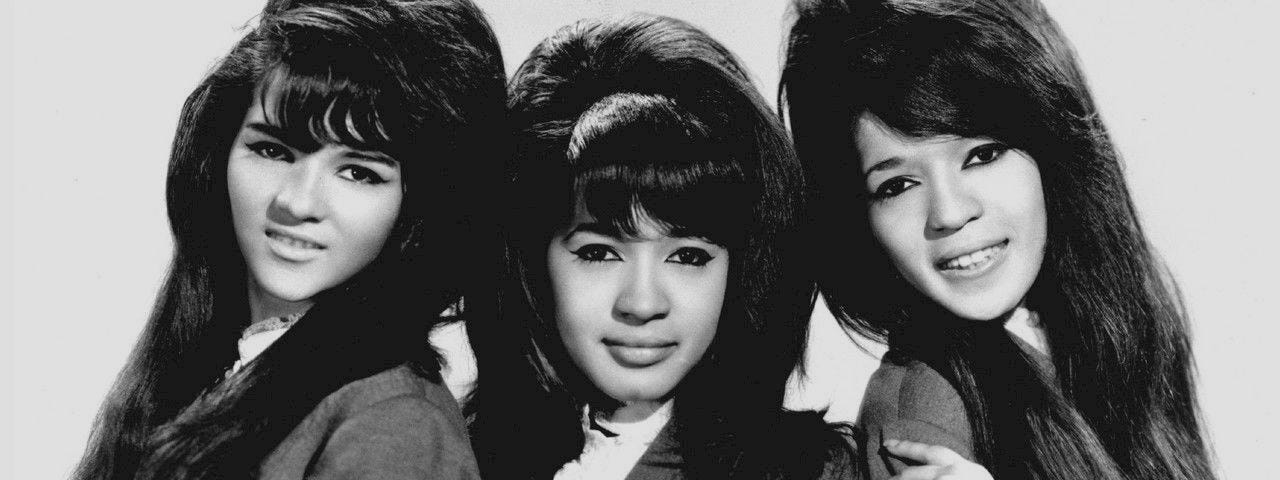 Black and white image of The Ronnettes in the 1960’s wearing matching suits and leaning into each other against a white background.