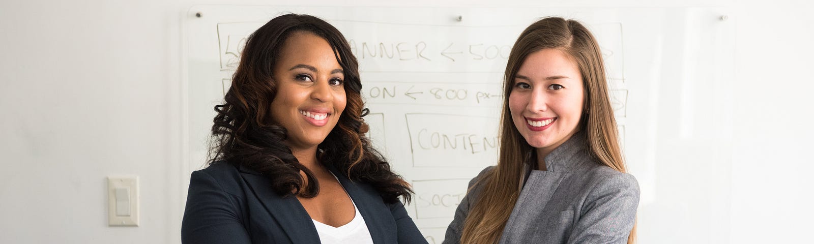Two females in work attire with crossed arms, smiling at the camera. White board in background.