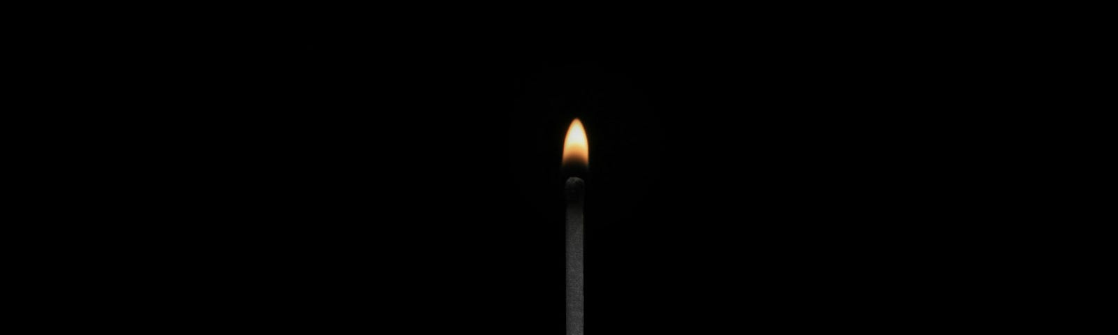 A black and white photo of a person holding up a lit match. The small fire is colored orange.