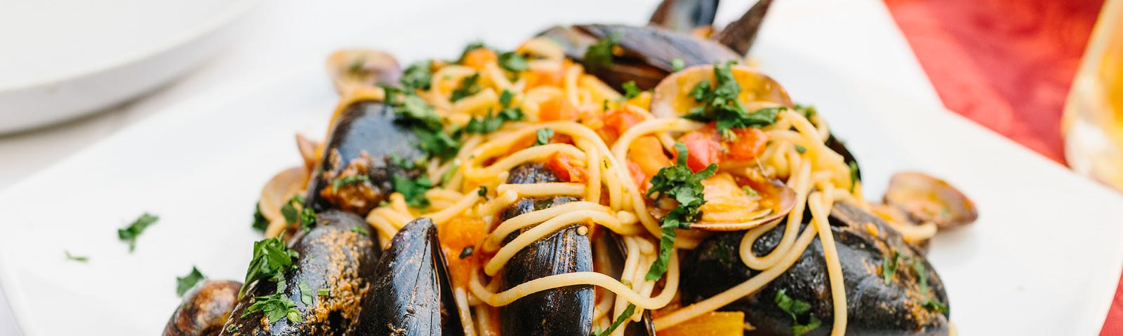 A square plate of spaghetti with fresh mussels and seafood on a table with a white tablecloth.