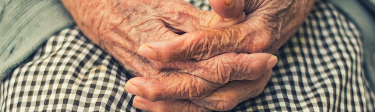 Wrinkled hands, folded, on a plaid skirt, of what seems like an elderly woman