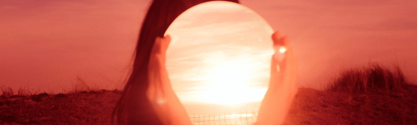 A woman holds a mirror over her face that reflects the bright light of the vivid red sunset.