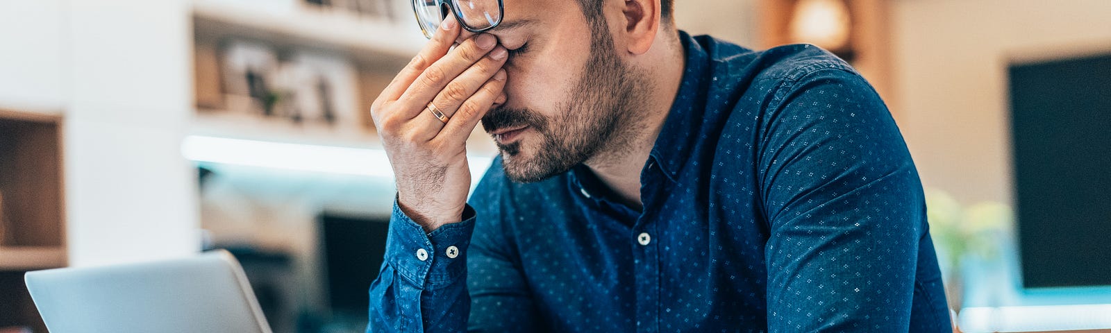 This image is a photograph of a writer at his desk with a laptop in front of him with eyes closed and  pinching the bridge of his nose with the fingers of his right hand in evident sadness, perhaps at what he has just written.