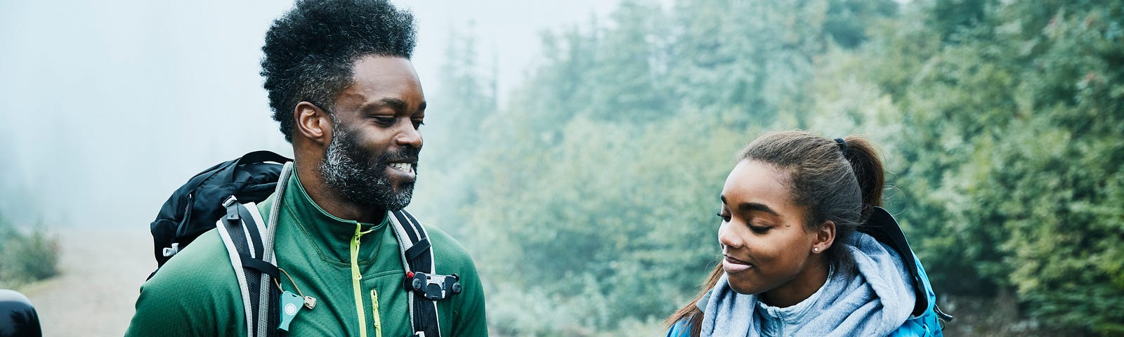 A smiling father and daughter stand and talk next to each other while preparing for early morning hike.