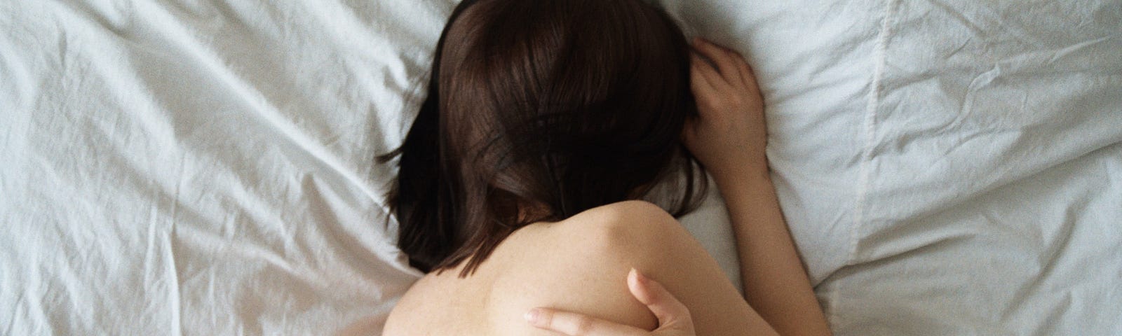You see the upper body of a brunette haired woman. She’s laying on her side on top of a bed with white sheets. Her hair is covering her face. You see her bare back. She has one of her arms wrapped around her upper torso.