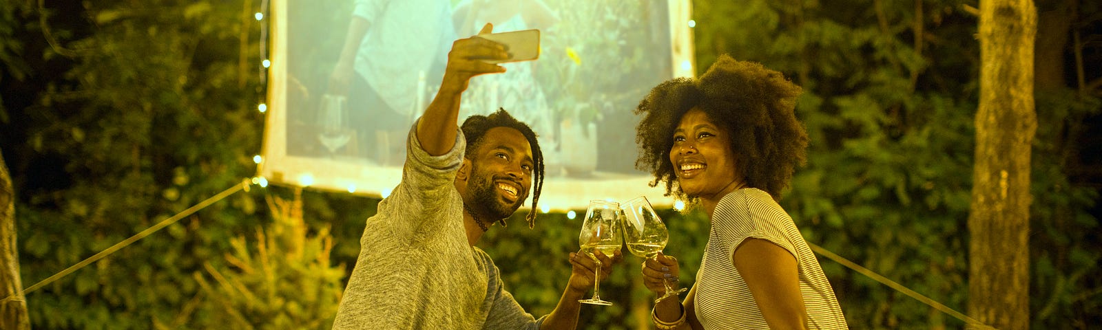 A Black couple take a selfie in their backyard, where they’ve set up a projector screen to watch a movie with hanging lights.