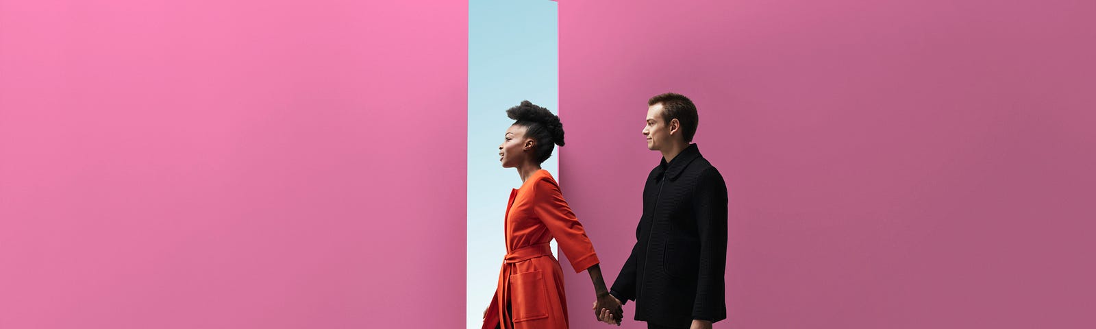 Interracial couple walking through a door in a pink wall, holding hands.