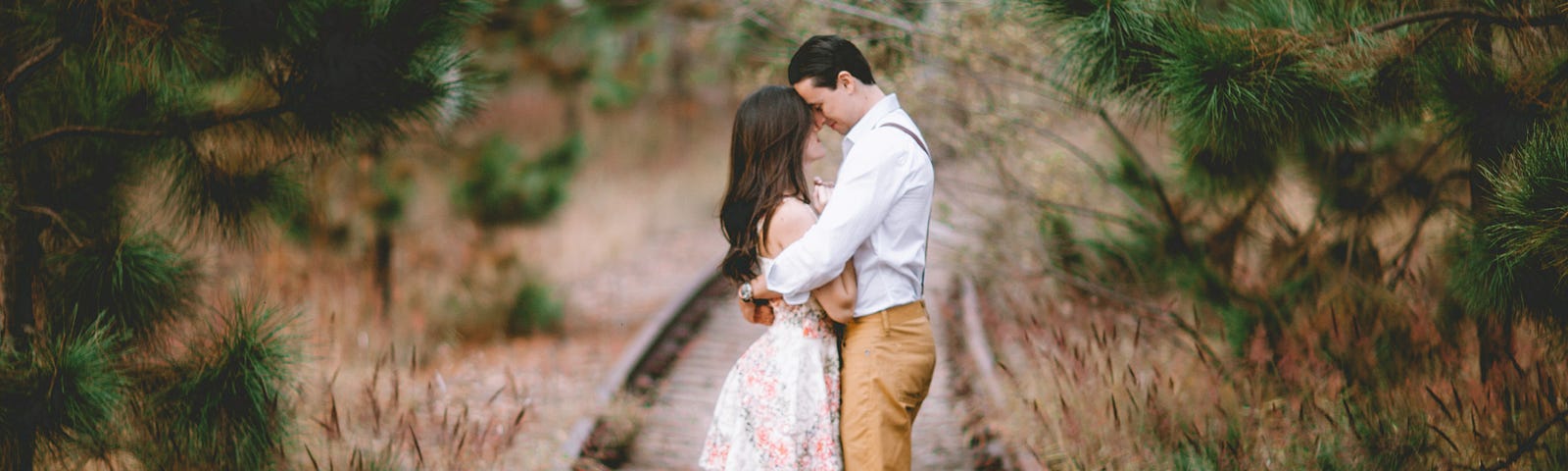 Couple standing together embraced on an old railroad track, surrounded by pine trees.