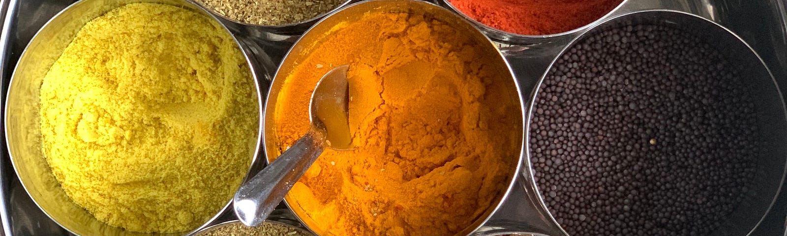 A large round high-walled metal dish containing 7 smaller metal containers filled with spices.