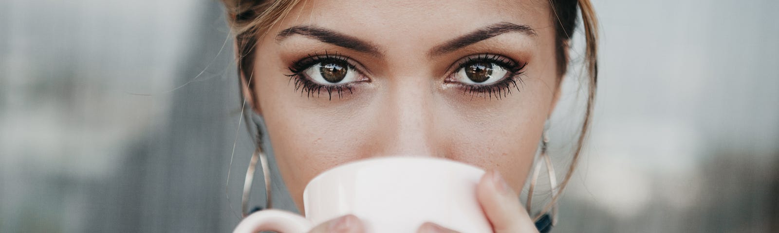 A Brunette with big brown eyes looking straight into the camera sipping coffee from a pink mug