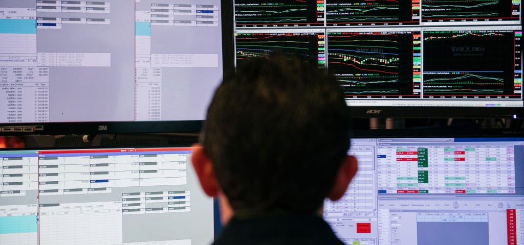 A photo that shows the back of a man’s head as he looks at stocks on four computer screens.