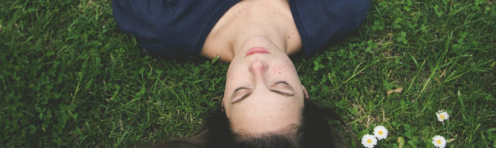 Young white woman in blue shirt looking calm with eyes closed, laying on her back in the grass