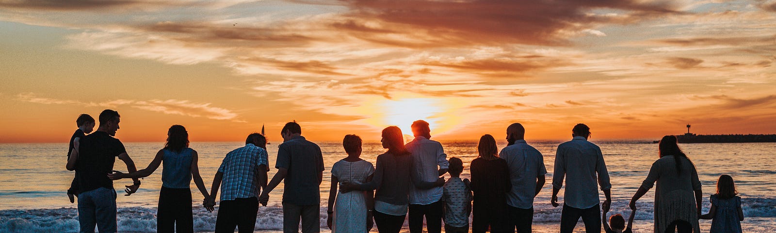 A large group of adults, children and babies holding hands and standing in a line on the beach, looking out to the horizon as the sun sets