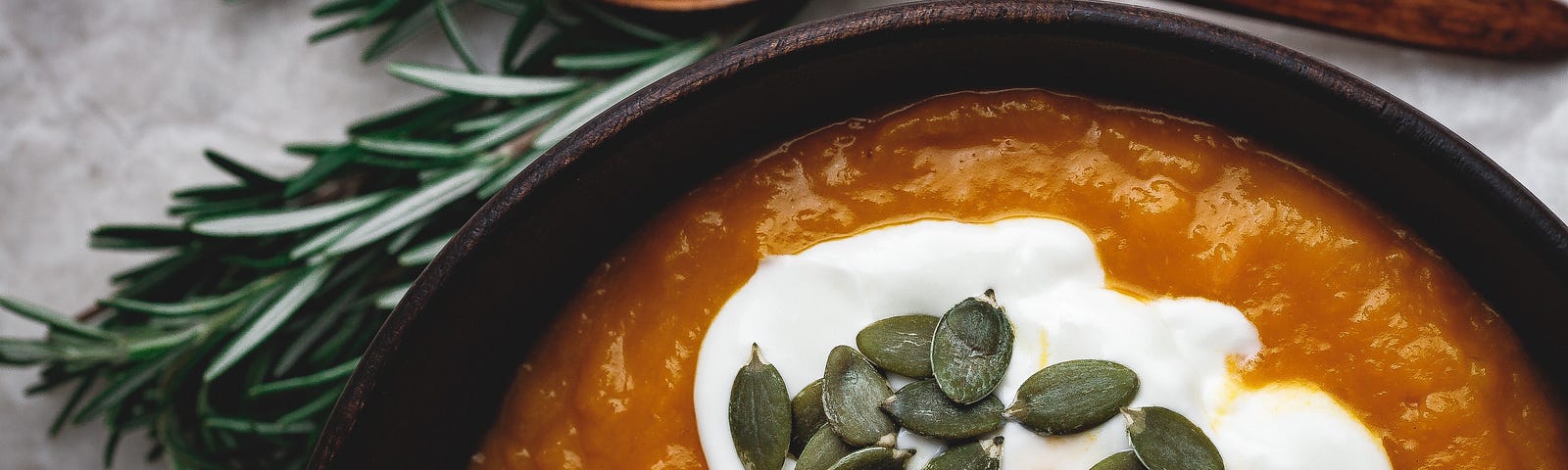 A bowl of soup with a fresh sprig of rosemary and a wooden spoon.