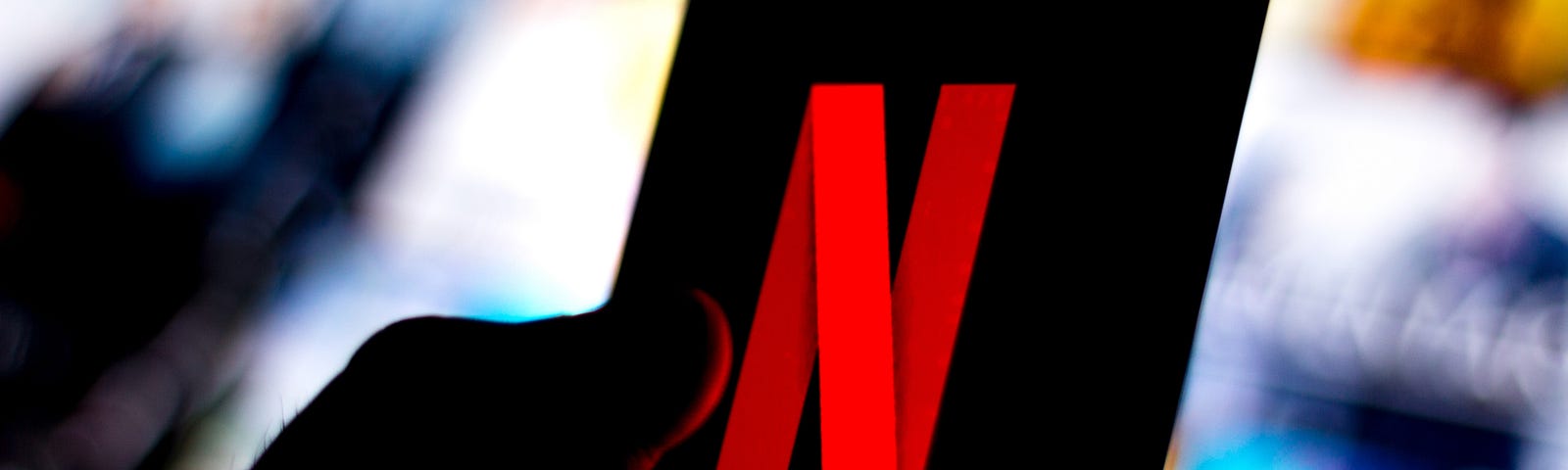 The Netflix logo is shown on a smartphone against a backdrop of blurred out Netflix show titles.