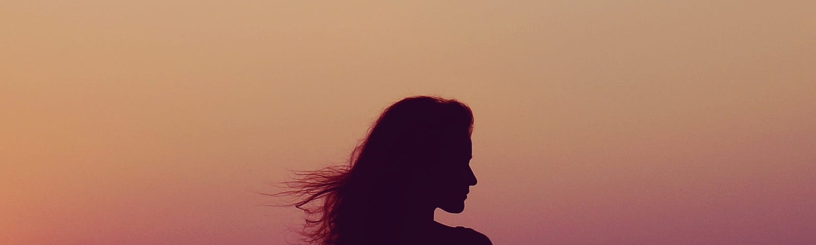 A person with long hair is silhouetted against a peach and purple sky at dusk.