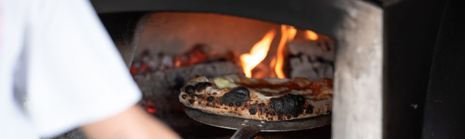 A person using a pizza peel to remove a pizza from an industrial metal wood-fired pizza oven.