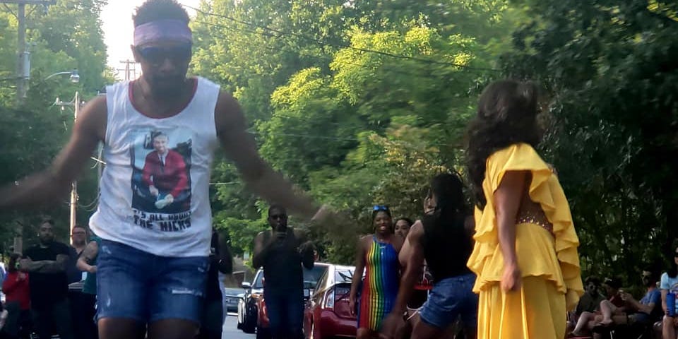 People dancing in summer clothing in the streets of a small town, one wearing an elaborate yellow dress.