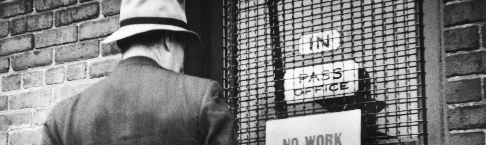 A black and white archival photo of a man standing in front of a door. The sign on the door says: ‘NO WORK. DON’T APPLY.’