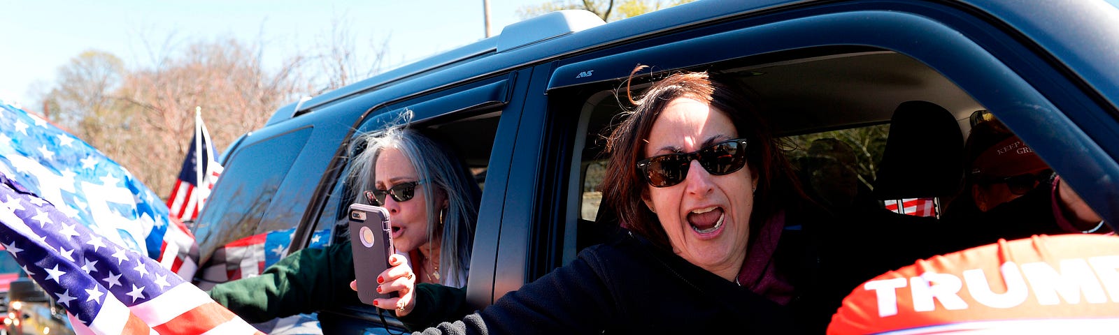 A photo of anti-shutdown protestors protesting from their car. Two white women hold US flags and “Trump 2020.”