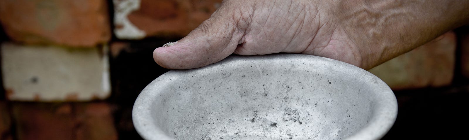 A photo of an old man holding out an empty bowl against a brick wall.