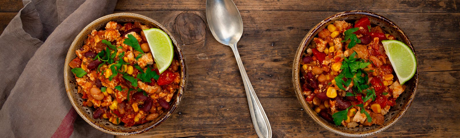 2 bowls of bean chili with a lime slice each, a cloth napkins, and a spoon between them, all on a rustic wooden surface.