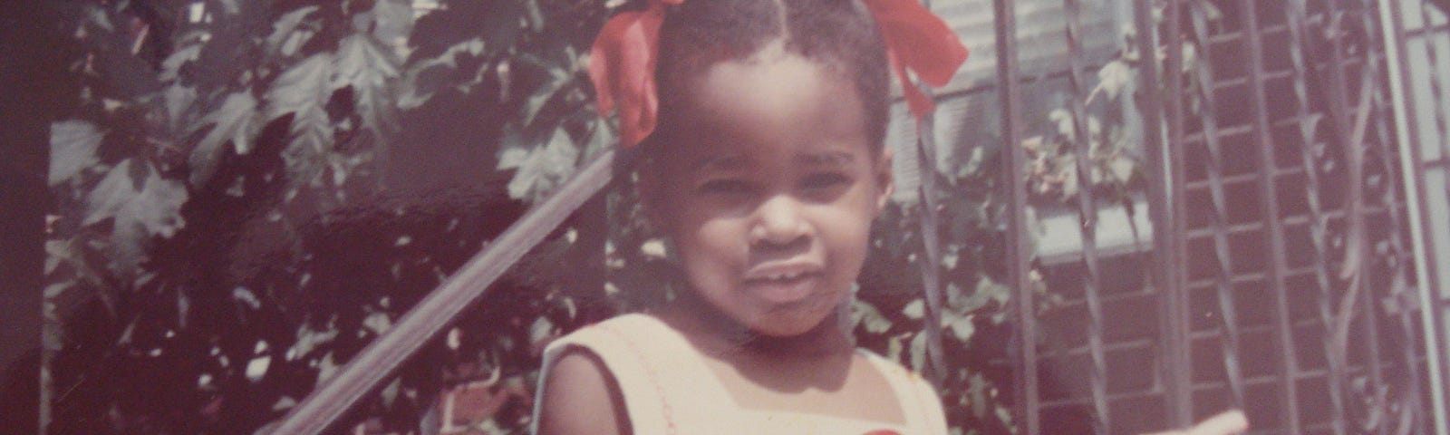A young, Black girl circa 1983 in a summer dress with two big, red ribbons in her hair.