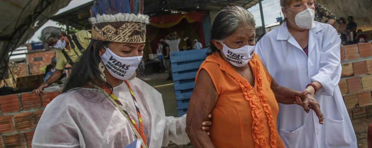 Two indigenous nurses with lab coats and face masks helping an elderly woman wearing a face mask and an orange dress walk.