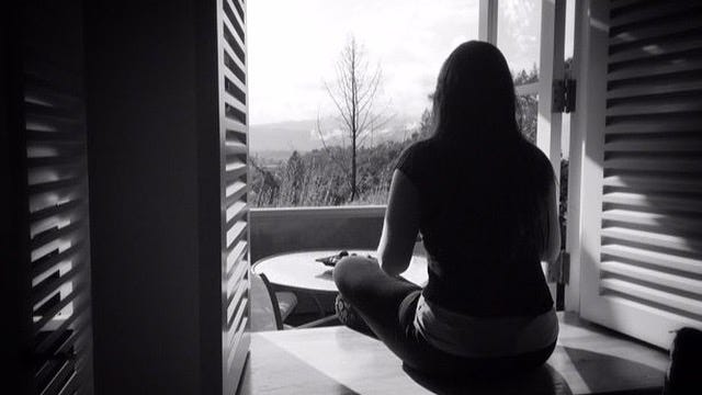 A black and white image shows a woman meditating on the sill of an open window with the hillside in the background.