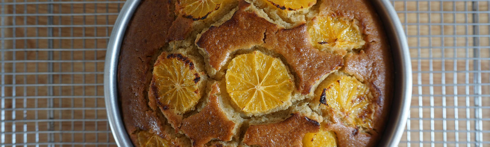 Overhead view of a cake still in its baking tin, resting on a wire rack. The cake is topped with 1 crosswise slice of orange in the center surrounded by 7 other slices.