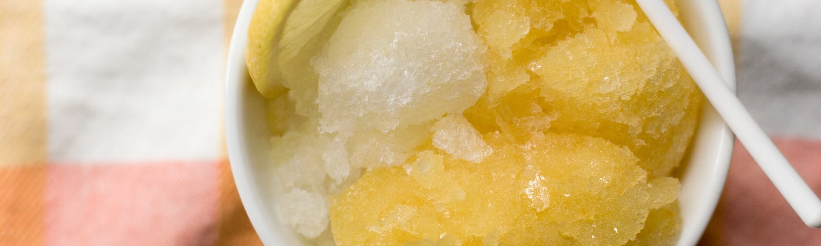 Overhead view of a small bowl of granita with a slice of lemon and a spoon on a background of pastel plaid fabric.