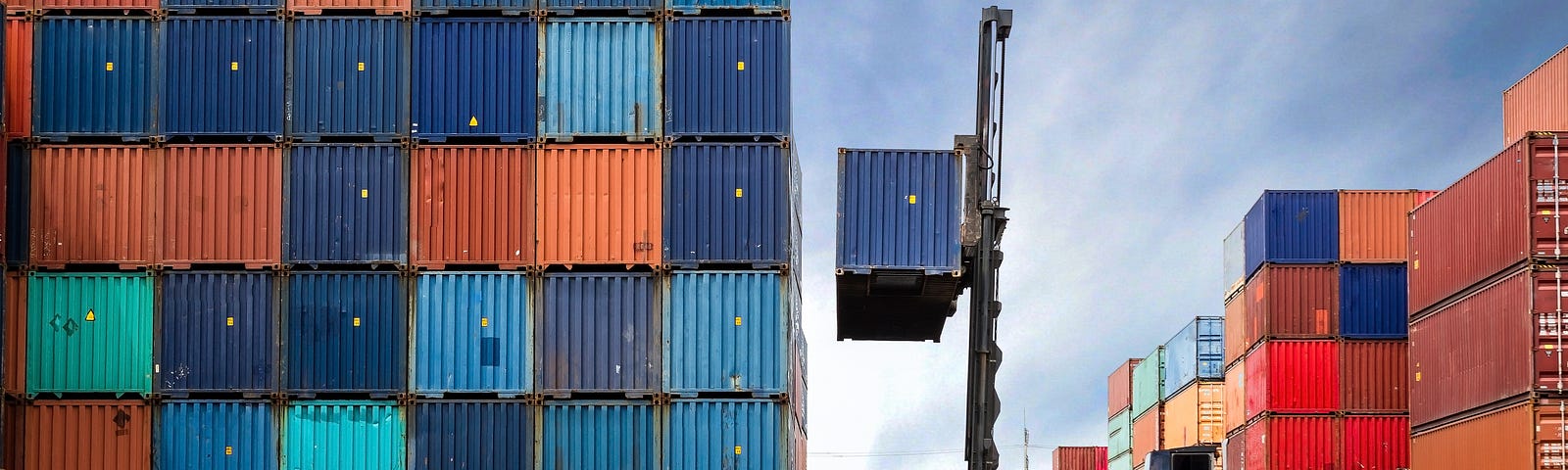 A crane lifting a shipping container next to a tall stack of other containers in a shipping yard.