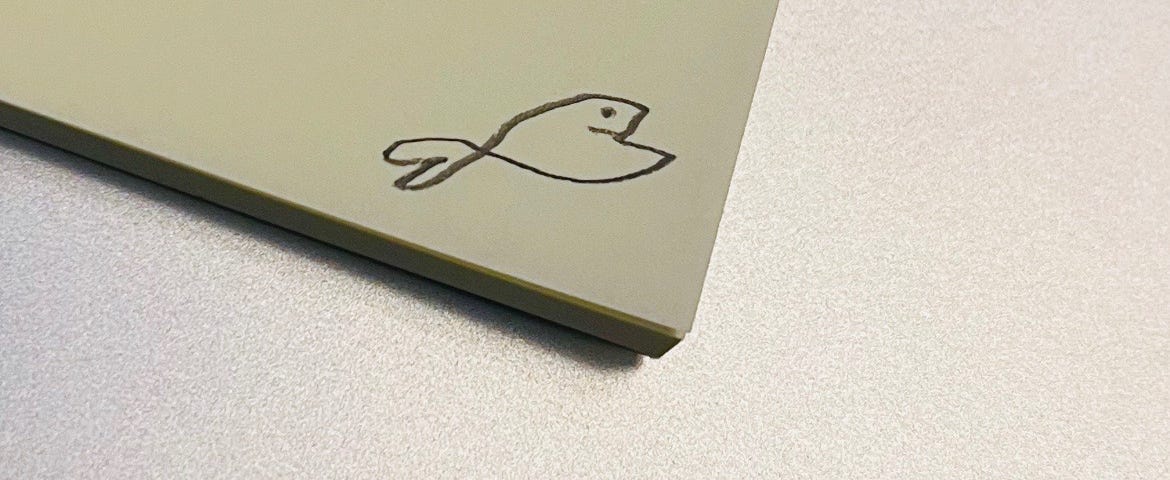 Tiny drawing of a goldfish on the corner of a yellow sticky pad, laying on the top of a silver MacBook.