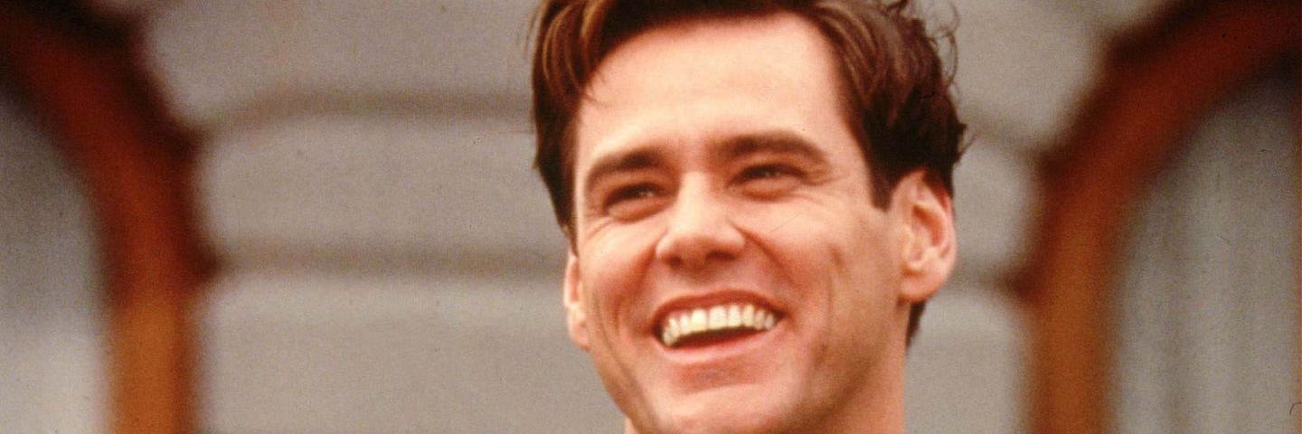 Jim Carrey in the movie The Truman Show, smiling on a bench wearing a biege patterned suit.