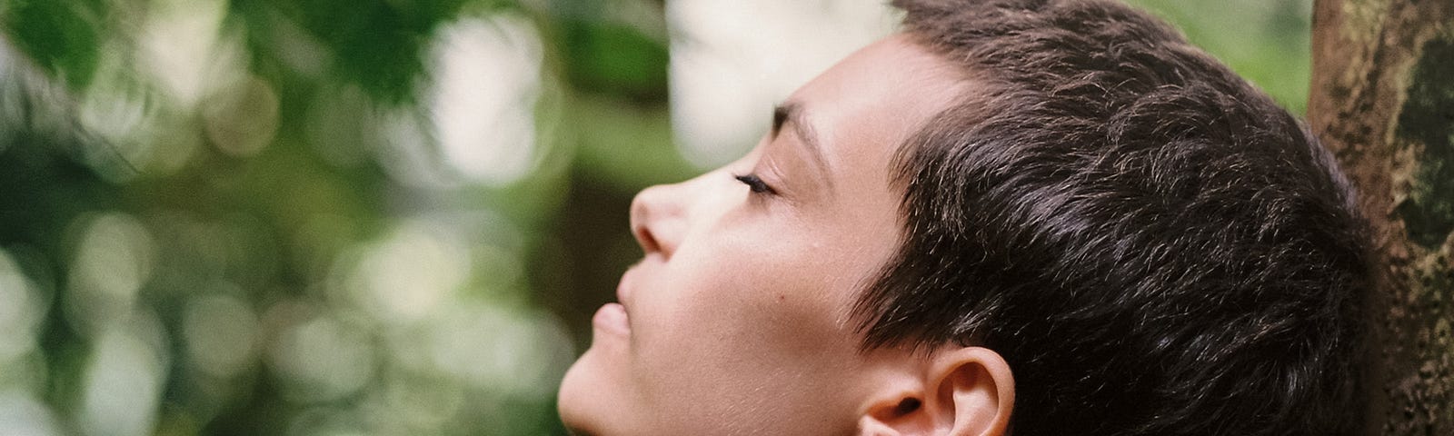 A person leans back against a tree with their eyes closed.