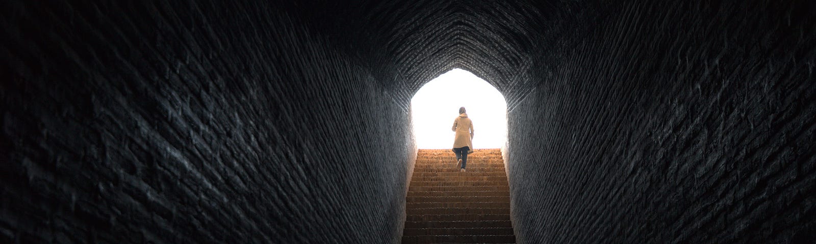 A lone woman walks with confidence up stairs coming out from dark tunnel into bright light.