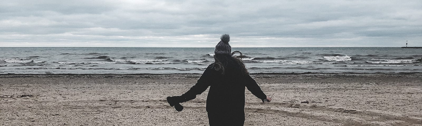 A woman runs away from the camera on a cold beach. She’s wearing a black coat, boots, and a purple beanie topped by a fuzzy ball.