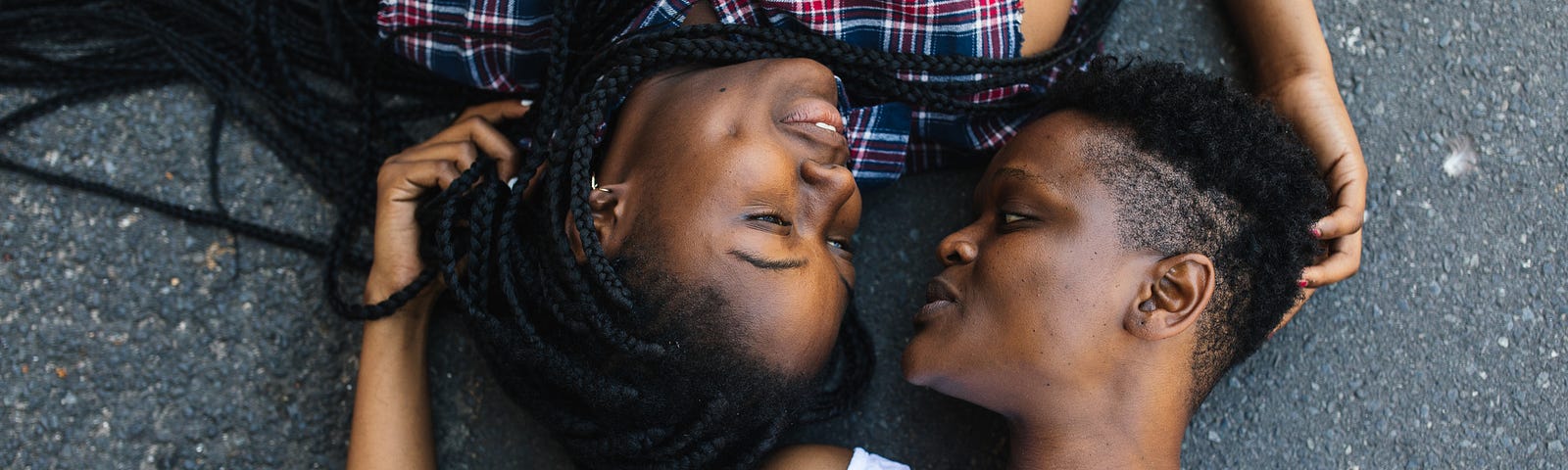 A photo of two black women lying down on pavement, looking at each other and touching each other’s hair.