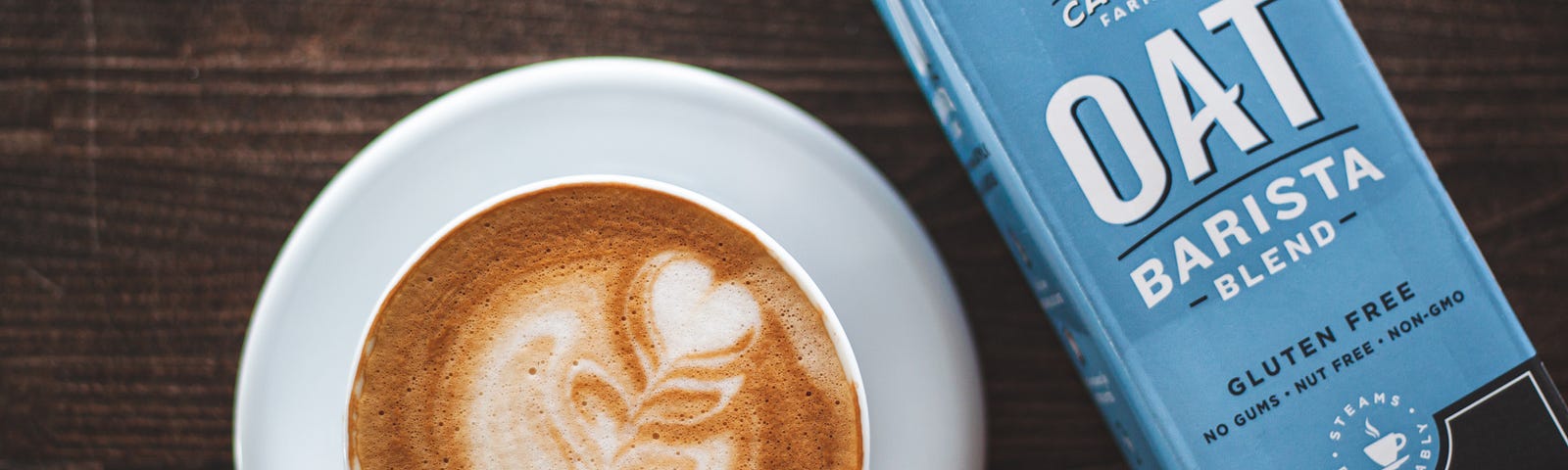 A latte with a flower shape in foam next to a carton of Califia Farms Barista Blend oat milk.