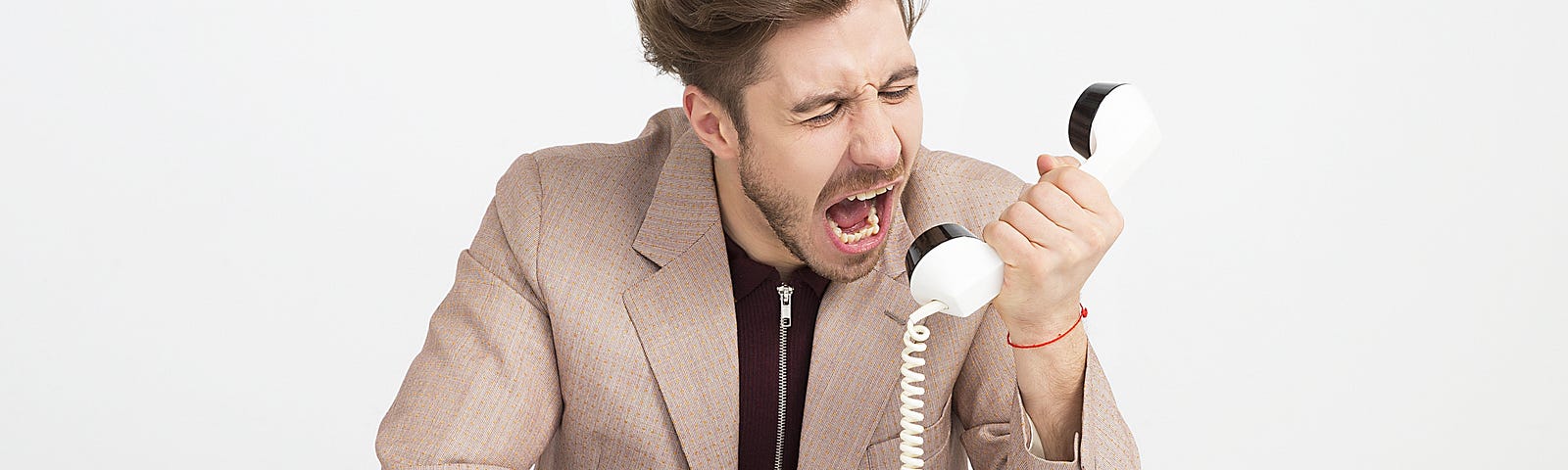Young man in beige blazer sitting at a desk screaming into an old fashion white phone