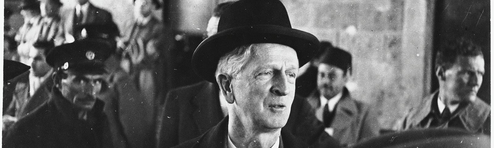 A man wearing an overcoat, vest, tie, and black hat looks off to the side of this black-and-white photograph. There are more men in business formal attire in the background. Over the man’s left shoulder, a man wearing a hat with a badge can be seen looking directly at the camera.