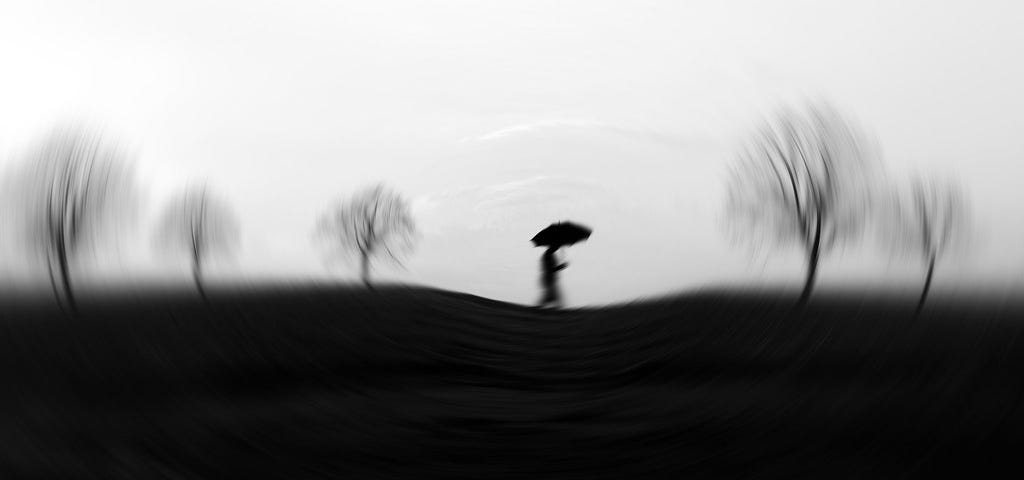 Blurry black and white image of a person walking holding an umbrella with some bare trees.