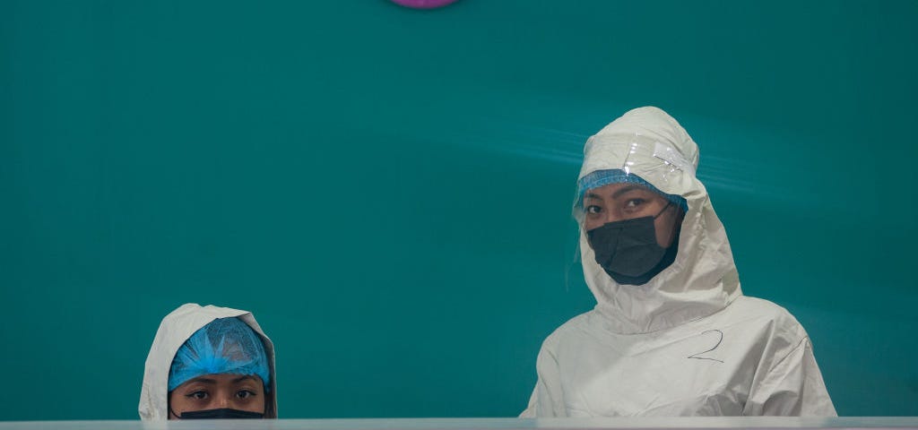 A photo of two healthcare workers wearing masks standing at a counter against a teal background.