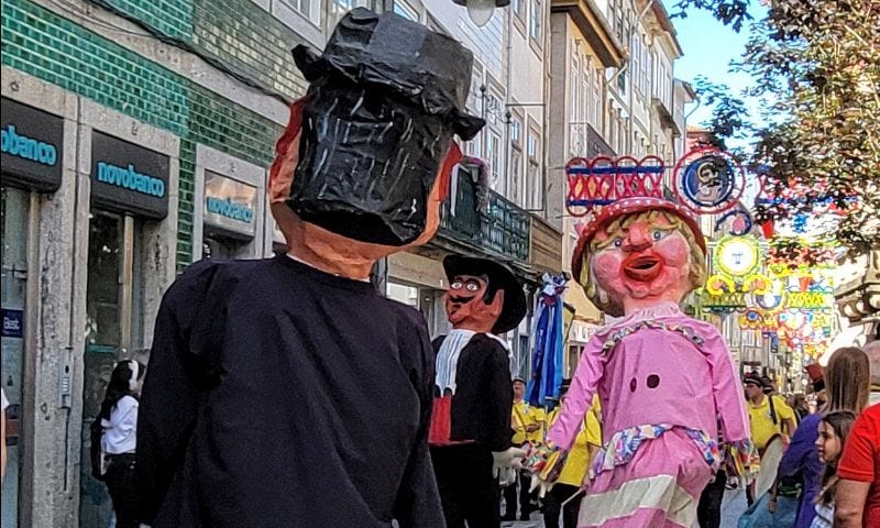 Three large puppets, two male puppets with black hair and black suits and one female with blond hair and a pink dress tower over the spectators that line the streets as they march in a parade.