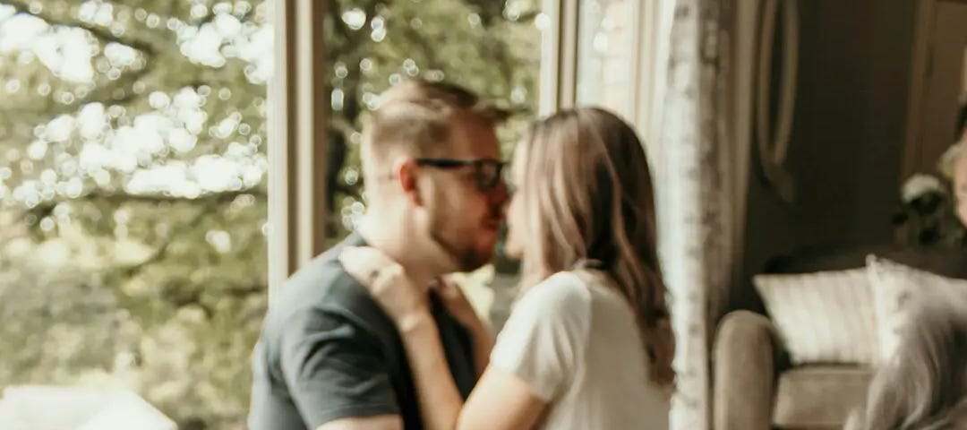 In the background a woman sits in the lap of a man, kissing him. In the foreground is a cat.