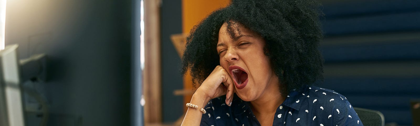 A young woman yawning at her desk while looking at her computer.