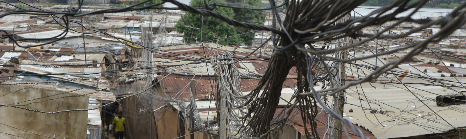 People walk under electric wires connected above roofs in a district of Abidjan, Côte d’Ivoire on June 1, 2019.