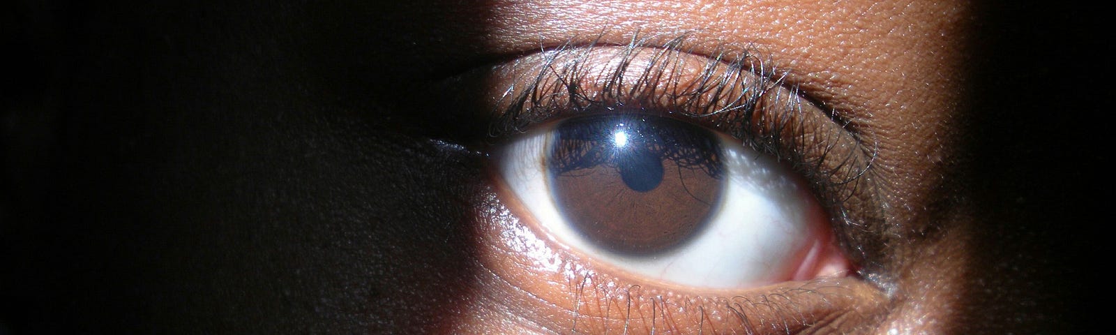 Closeup of a Black woman’s eye, with light shining in a strip where her eyes is. Rest of face obscured by shadow.