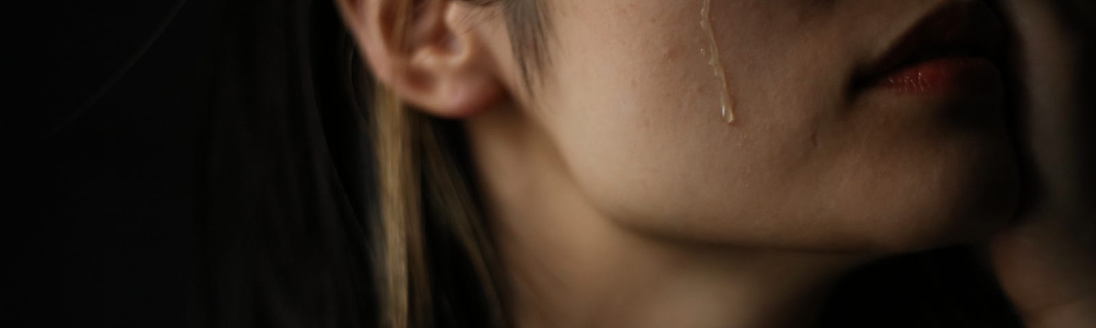 Moody shot of a person with long hair and a tear rolling down their cheek. Their eyes are out of frame.