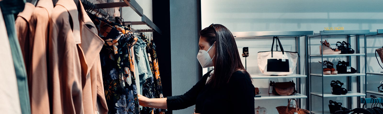 A person with long hair, wearing a mask and carrying one shopping bag, flips through clothes at a retail store.